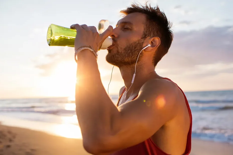 Uomo che beve acqua da bottiglia verde dopo allenamento sulla spiaggia al tramonto per idratarsi rapidamente.
