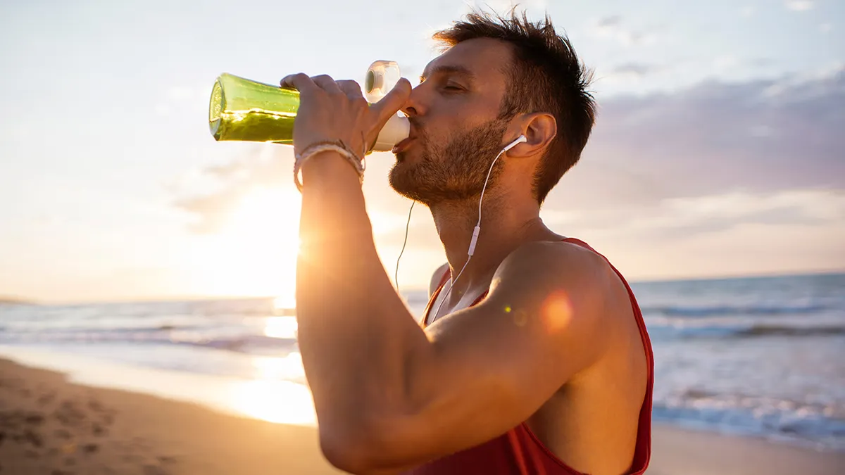 Uomo che beve acqua da bottiglia verde dopo allenamento sulla spiaggia al tramonto per idratarsi rapidamente.