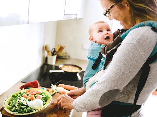 una mamma davanti alla cucina con il suo bambino in braccio con davanti un piatto di verdure da mangiare durante l'allattamento.