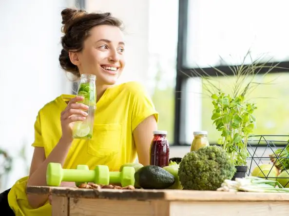 Una donna che siede ad un tavolo con pesi e cibi sani mentre stringe un bicchiere di succo di frutto ad indicare la dieta di mantenimento dopo la terapia farmacologica dimagrante.